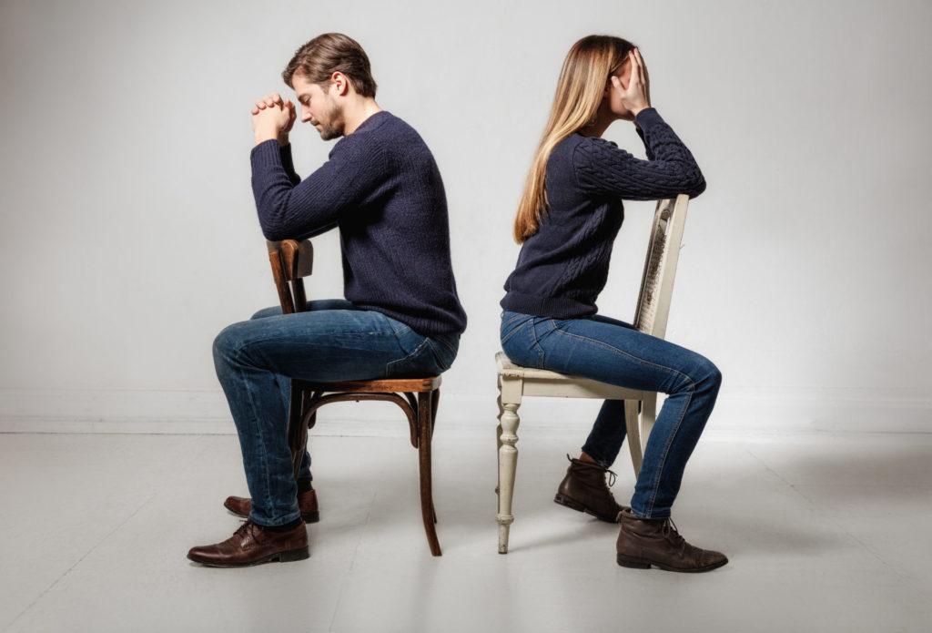 Seitenansicht eines depressiven Paares, das Rücken an Rücken auf Stühlen vor grauem Hintergrund sitzt und von Paartherapie profitieren könnte.